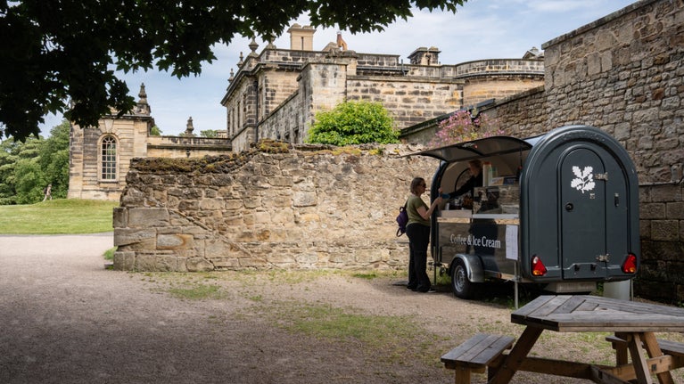 A long black horse trailer which has been converted into a catering kiosk sits in front of a stone wall. Behind the wall is the corner of a grand stone building. In the foreground there is a picnic bench. A woman is standing at the kiosk, which has 'Coffee & Ice Cream' written on the front